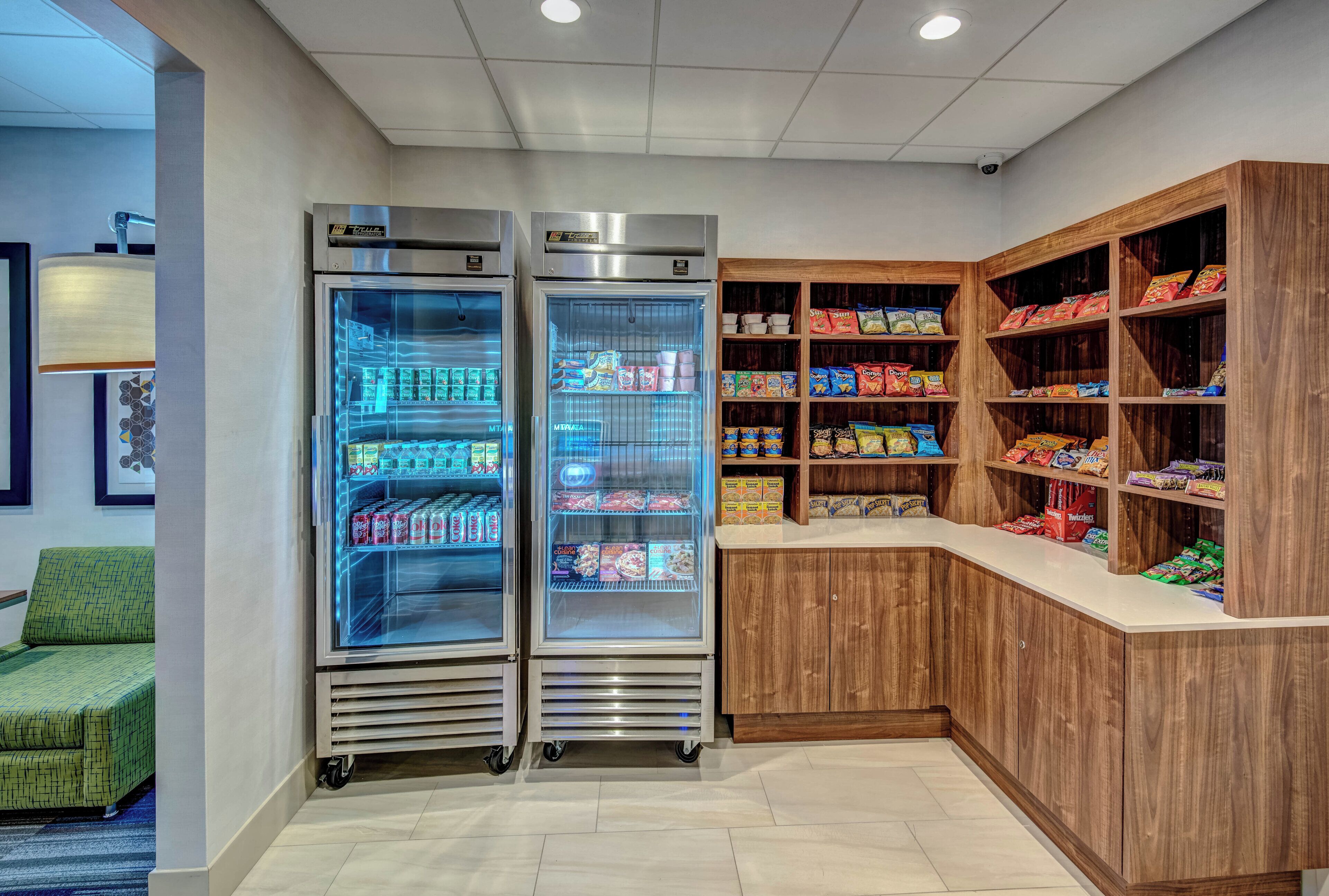 Rincón de una habitación de hotel con dos refrigeradores llenos de bebidas, una estantería de madera con aperitivos y un sofá verde estampado.