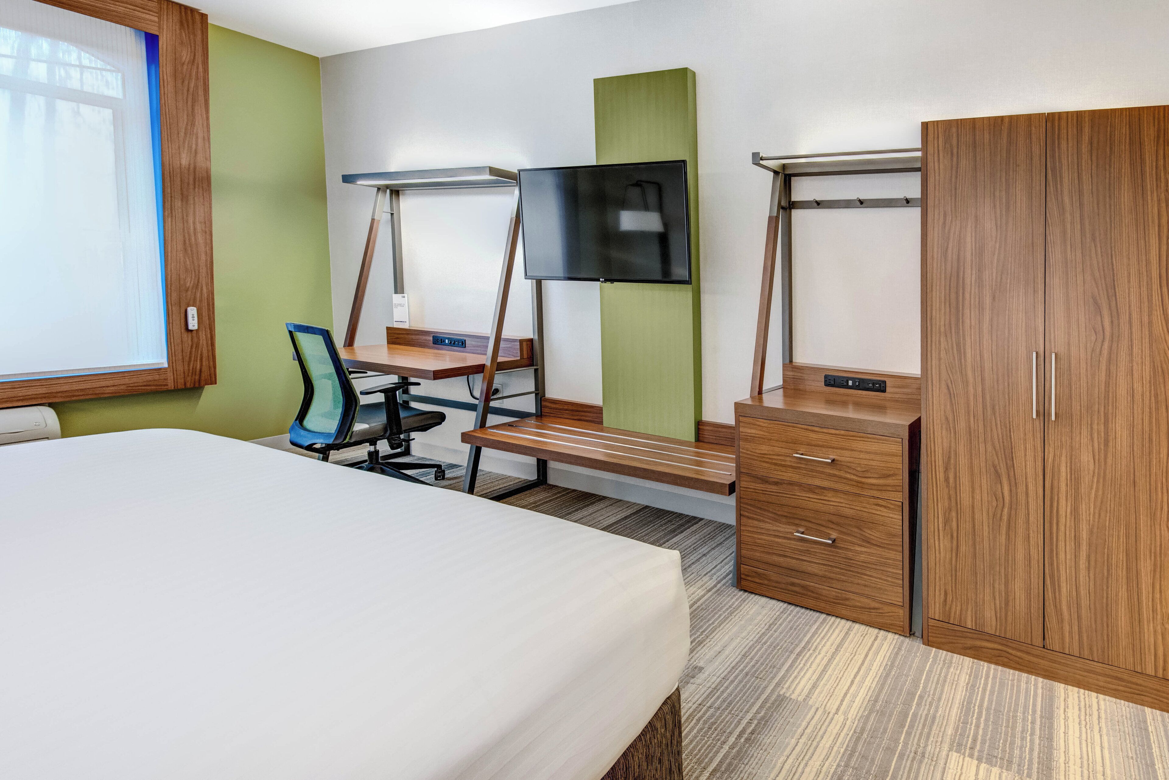 A hotel room with a bed, desk, chair, TV, wardrobe, and a window.