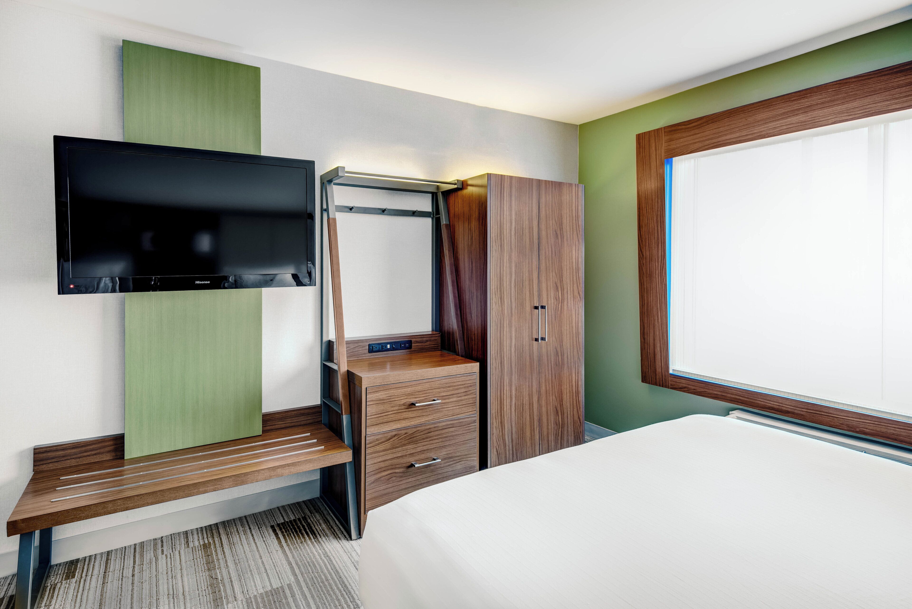 A hotel room with a bed, a TV mounted on the wall, a wooden dresser with drawers, and a wardrobe.