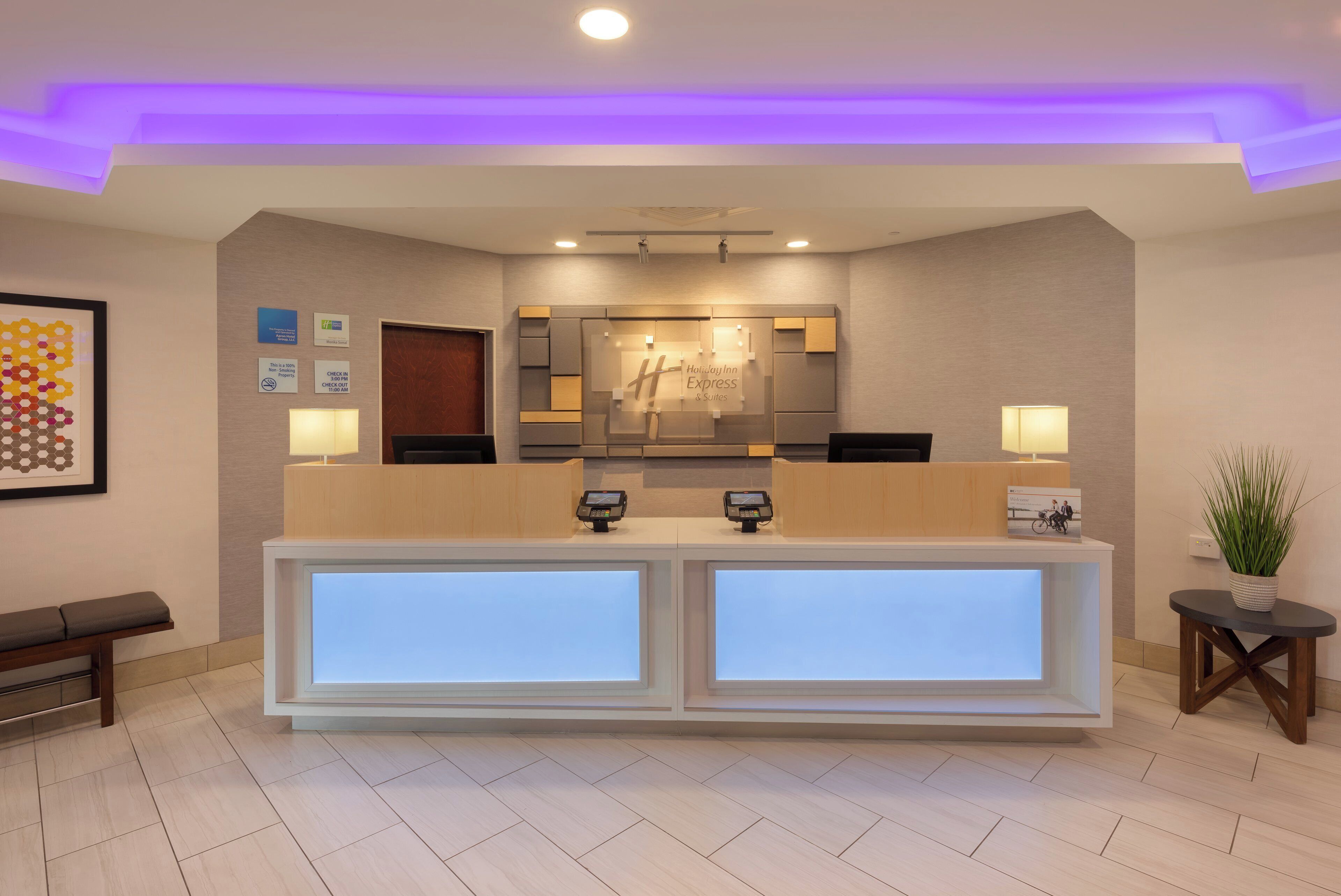 Zona de recepción de hotel con diseño moderno, que incluye un mostrador de recepción con pantallas digitales, un mostrador de registro y un mostrador con un espejo grande.
