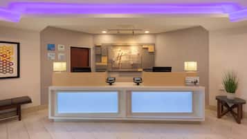 Zona de recepción de hotel con diseño moderno, que incluye un mostrador de recepción con pantallas digitales, un mostrador de registro y un mostrador con un espejo grande.