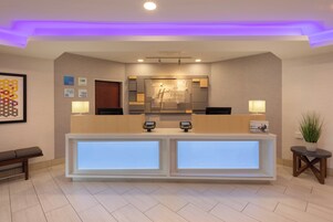 Zona de recepción de hotel con diseño moderno, que incluye un mostrador de recepción con pantallas digitales, un mostrador de registro y un mostrador con un espejo grande.