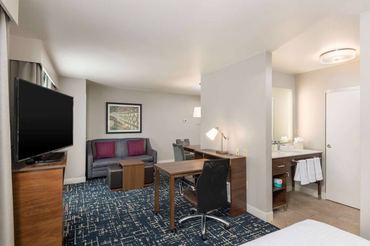 Habitación de hotel con televisión de pantalla plana, sofá, escritorio con silla, baño con lavamanos y espejo, y alfombra estampada.
