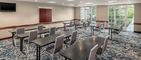 Une salle de conférence dotée de plusieurs tables et chaises, d’un téléviseur à écran plat fixé au mur et de grandes fenêtres laissant entrer la lumière naturelle.