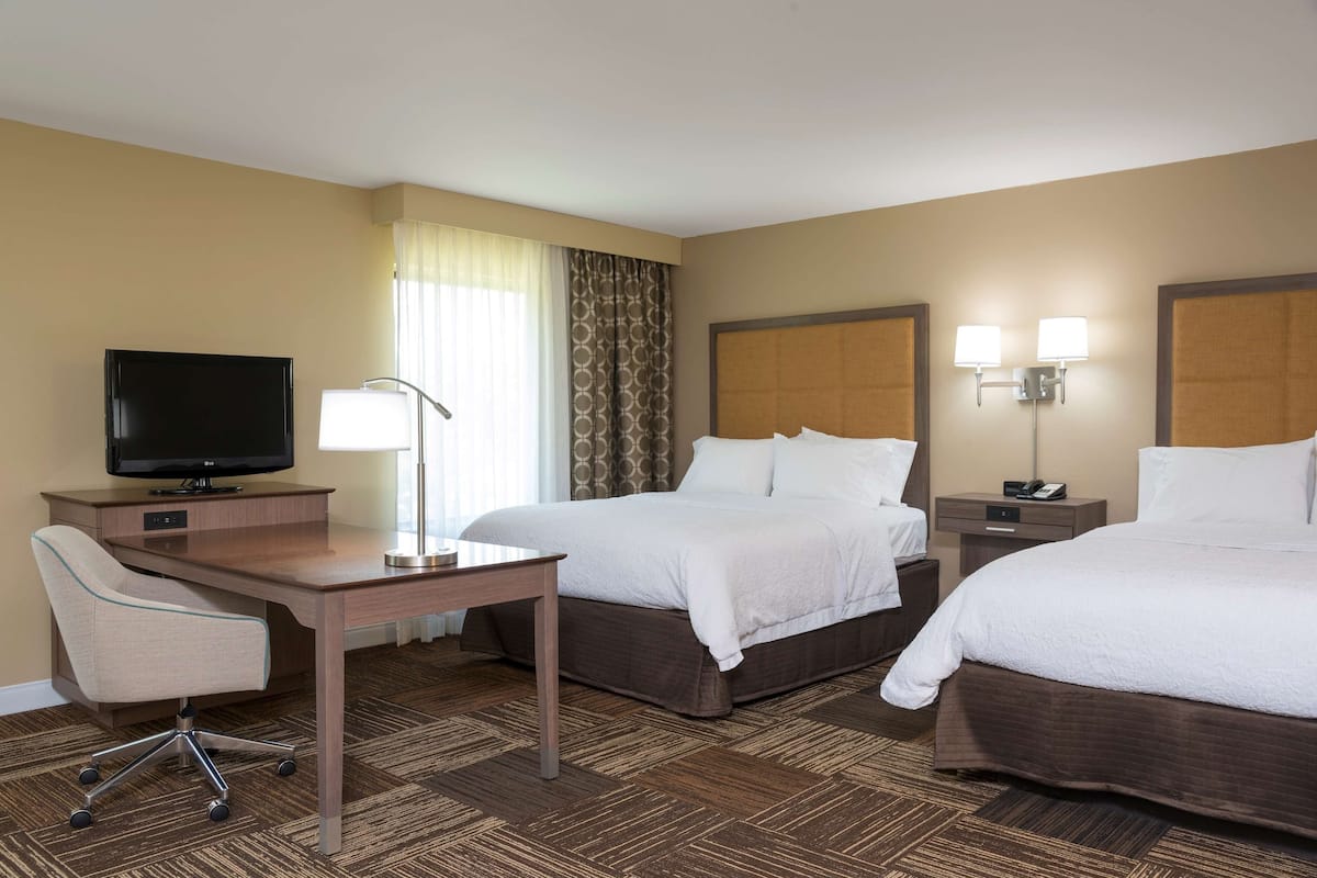 A hotel room with two beds, a desk with a television, a chair, and a lamp.