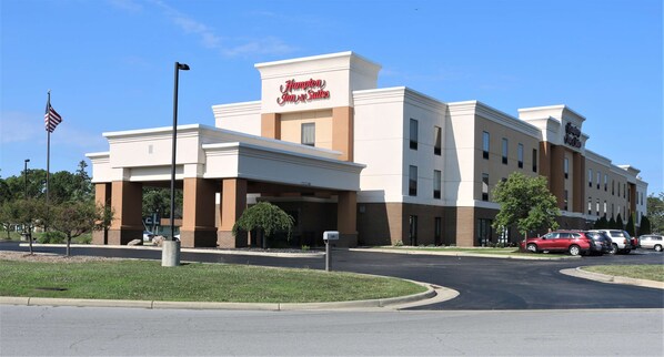 A Hampton Inn Suites building with a modern architectural design, featuring a prominent entrance and a parking area with several cars.