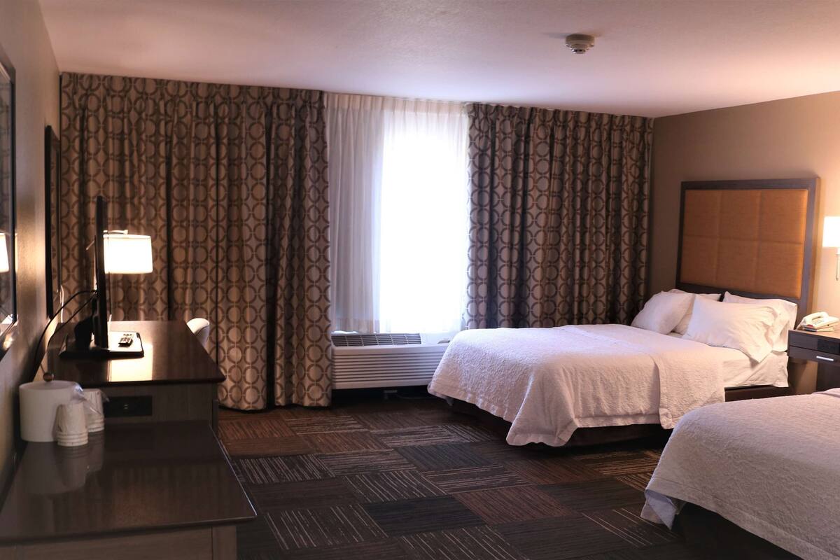 A hotel room with two beds, a desk with a lamp, a telephone, and a window with curtains.