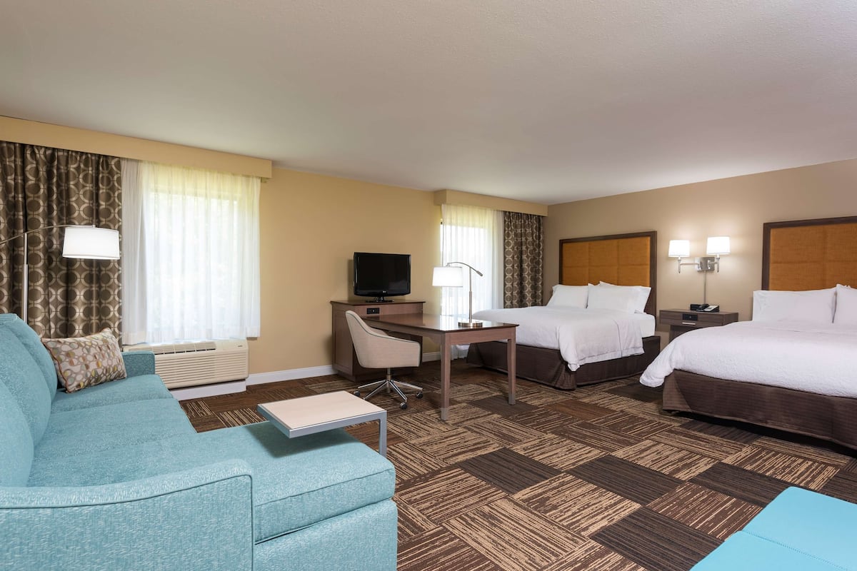 A hotel room with a sofa, a desk, a TV, two beds, and a window with curtains.