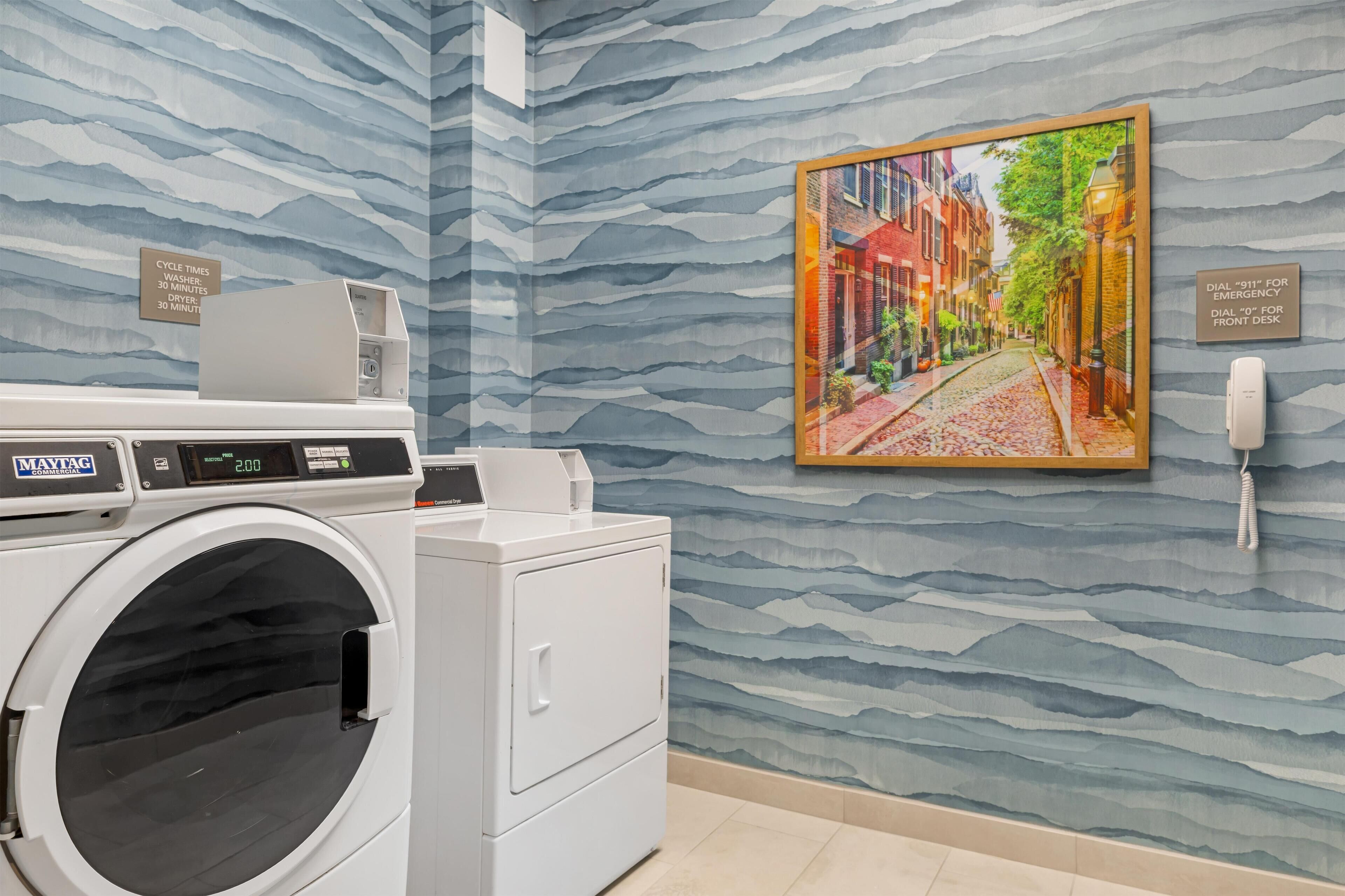 A modern laundry room with a front-loading washer and dryer, a wall-mounted phone, and a framed cityscape painting.