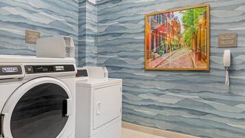 A modern laundry room with a front-loading washer and dryer, a wall-mounted phone, and a framed cityscape painting.