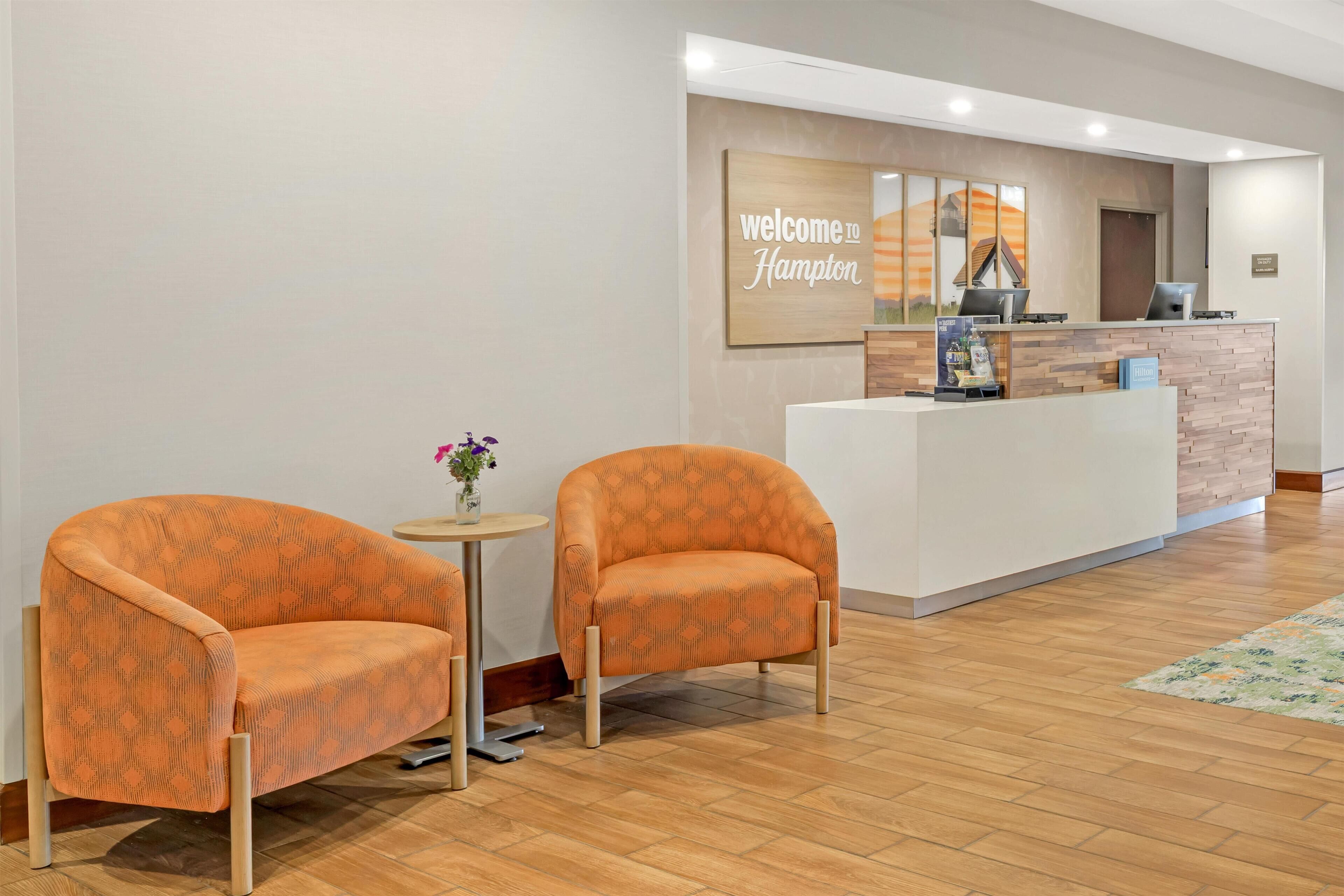 A reception area with two orange armchairs, a small round table with a vase of flowers, and a reception desk with a 'Welcome to Hampton' sign.