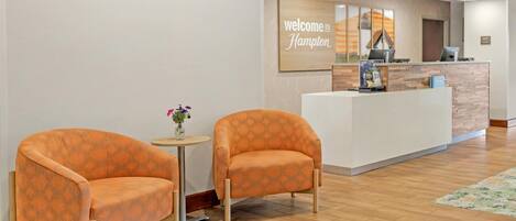 A reception area with two orange armchairs, a small round table with a vase of flowers, and a reception desk with a 'Welcome to Hampton' sign.