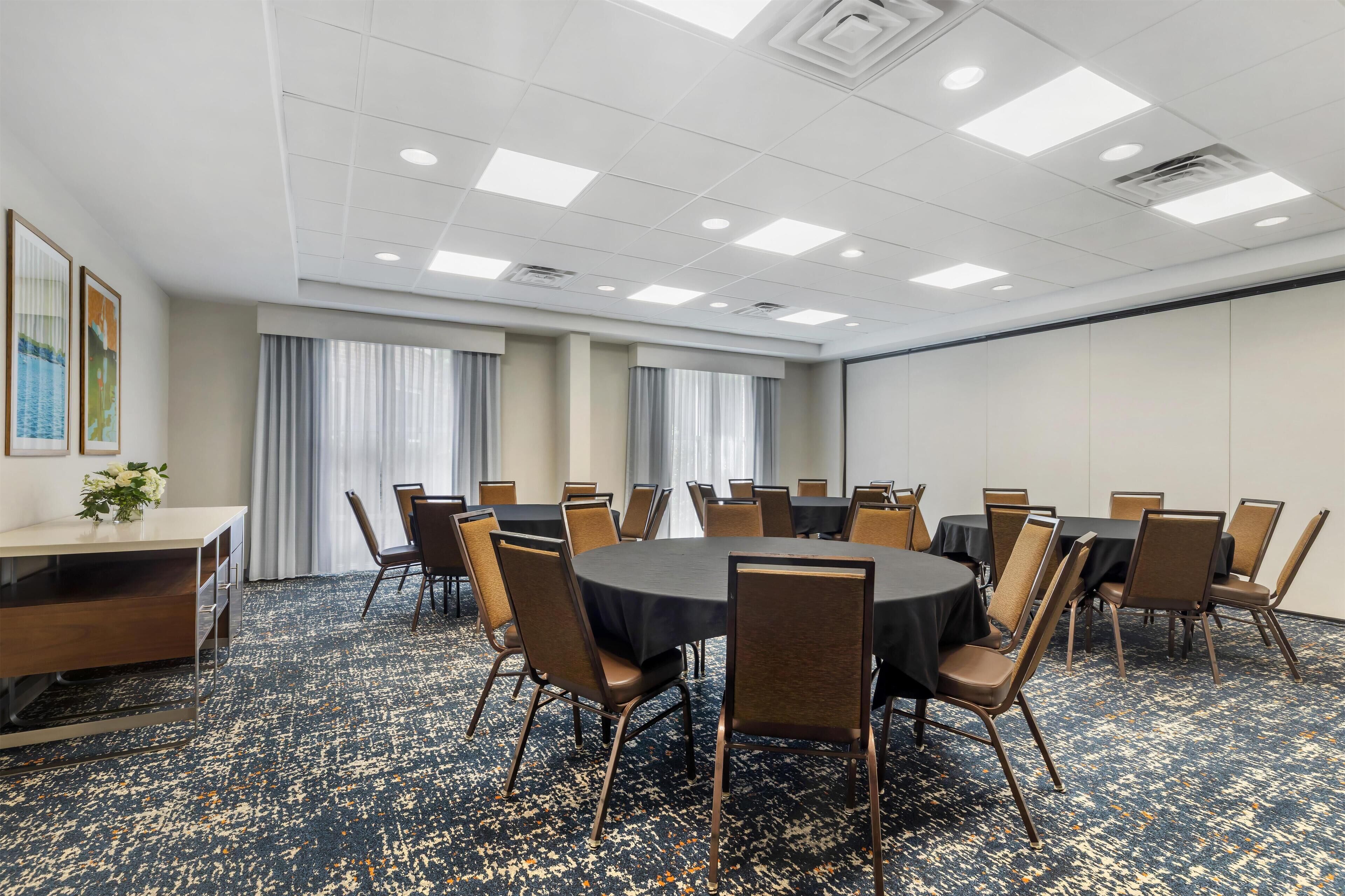 A conference room with a round table, chairs, a desk, and a large window with curtains.