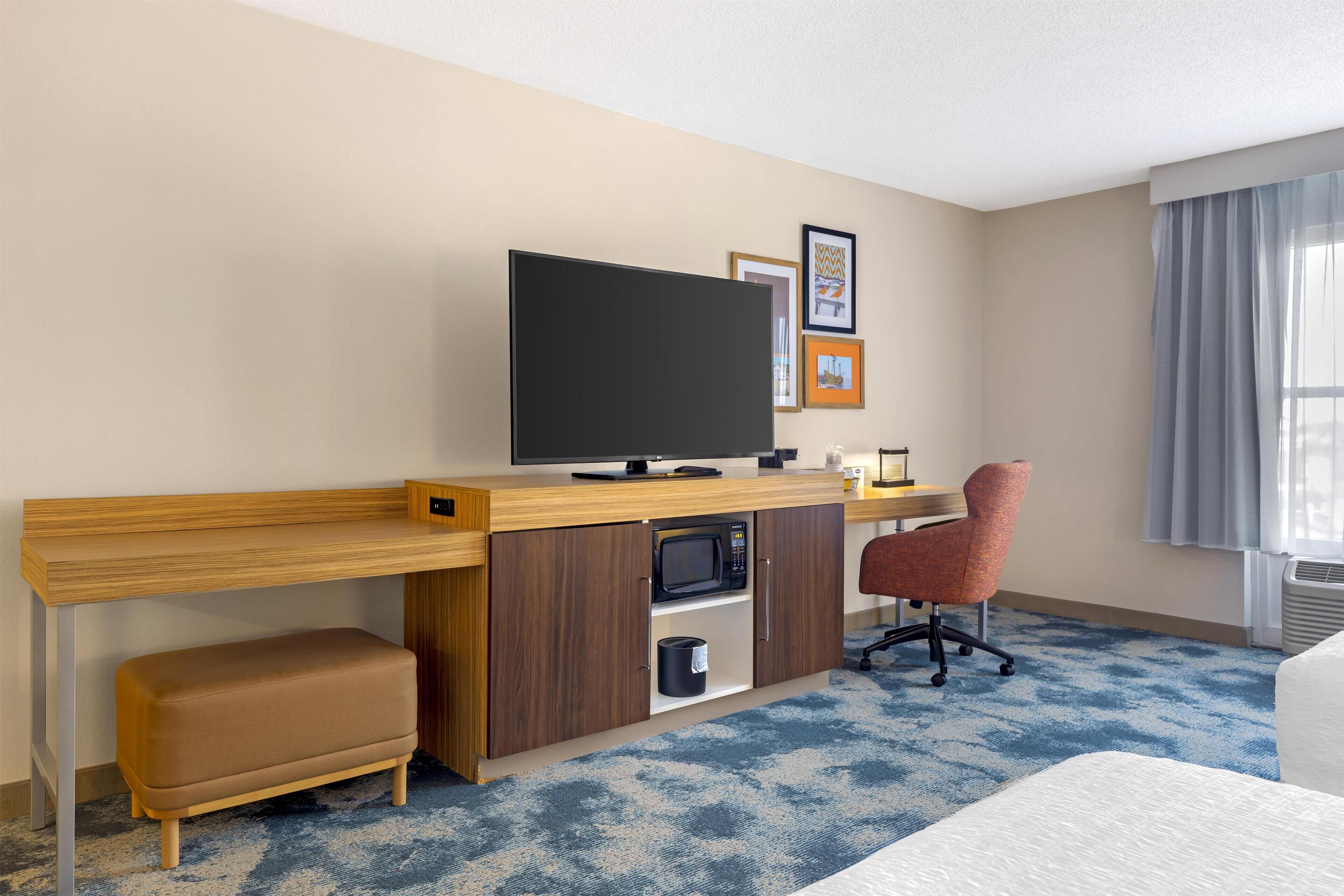 A hotel room with a wooden desk, a television, a microwave, a cushioned bench, a red office chair, and framed pictures on the wall.