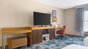 A hotel room with a wooden desk, a television, a microwave, a cushioned bench, a red office chair, and framed pictures on the wall.