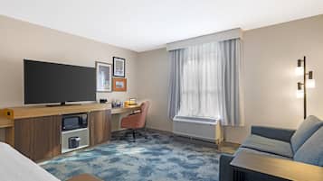 A hotel room with a bed, a desk with a computer, a television, a microwave, a chair, a sofa, and a large window with curtains.