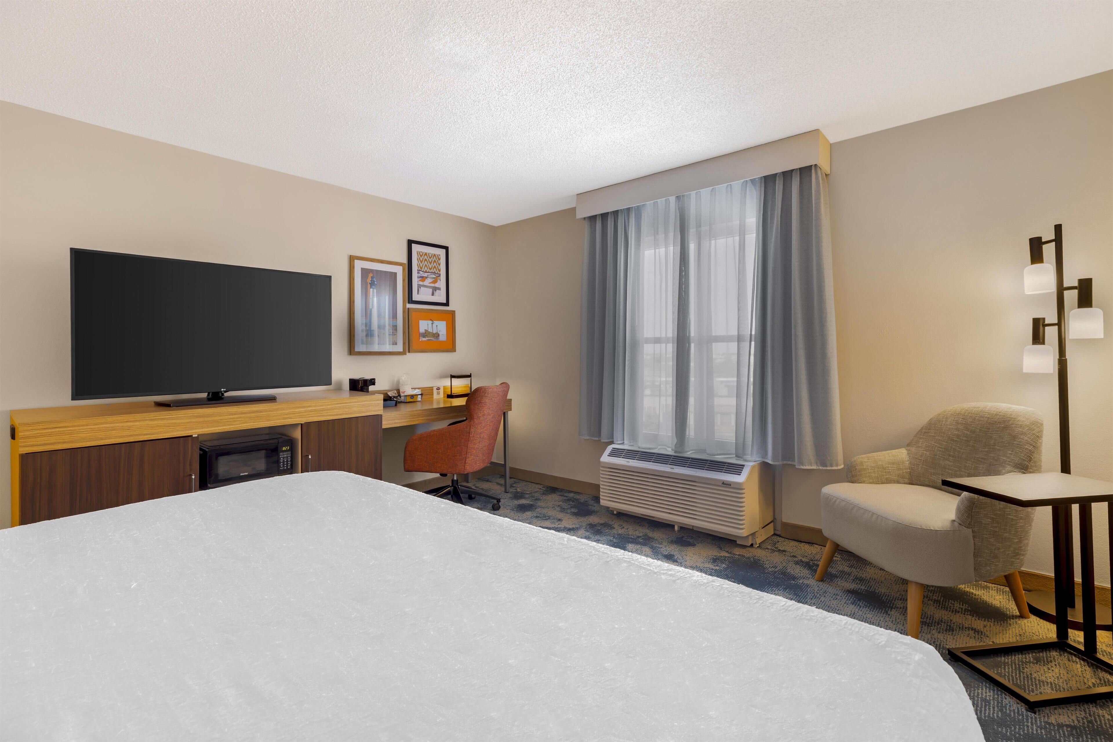 Habitación de hotel con una cama grande, un escritorio con televisión de pantalla plana, una silla, una mesita y una ventana con cortinas.