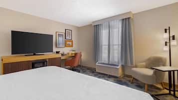 Habitación de hotel con una cama grande, un escritorio con televisión de pantalla plana, una silla, una mesita y una ventana con cortinas.