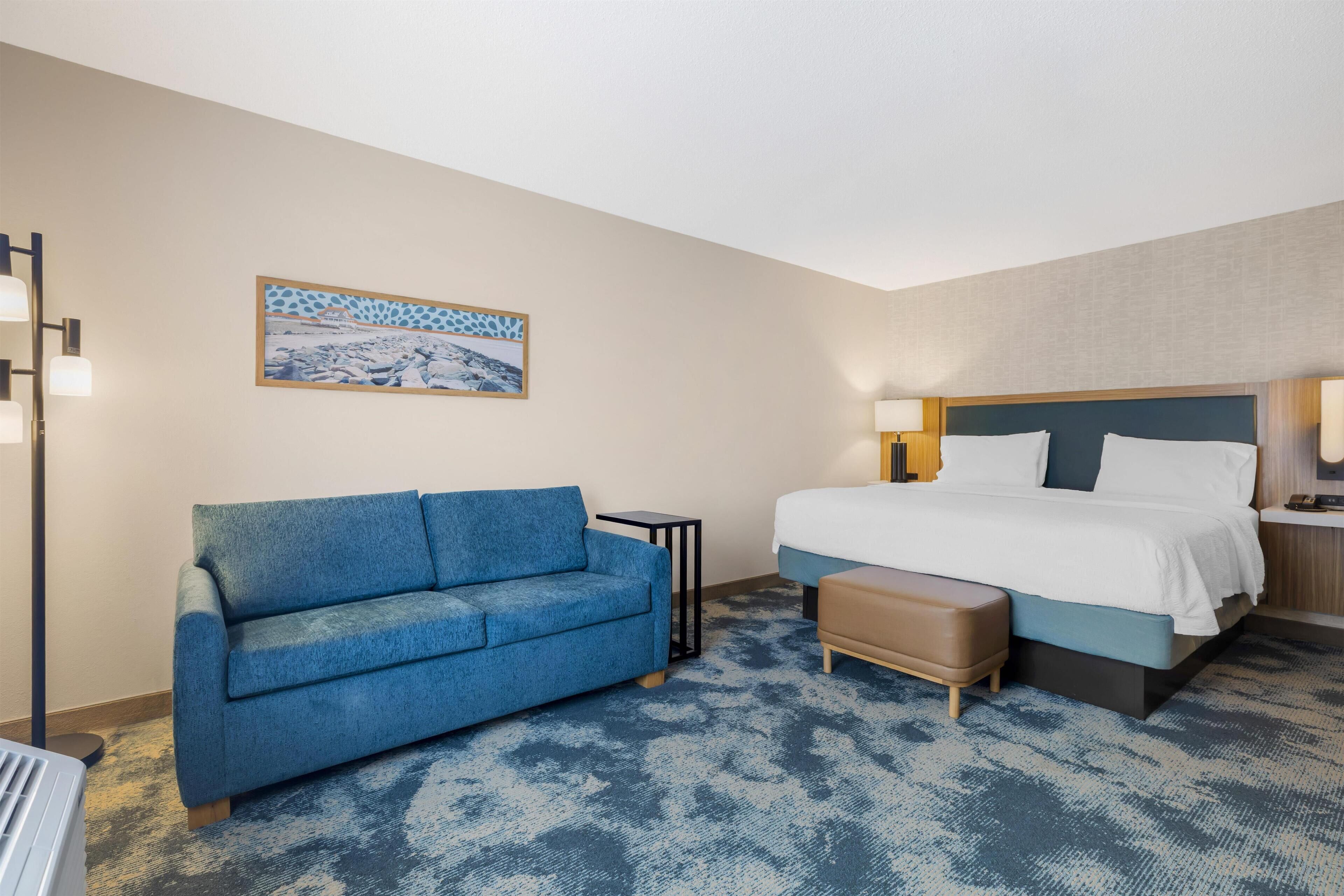 A hotel room with a bed, a blue sofa, a small table, and a painting on the wall.
