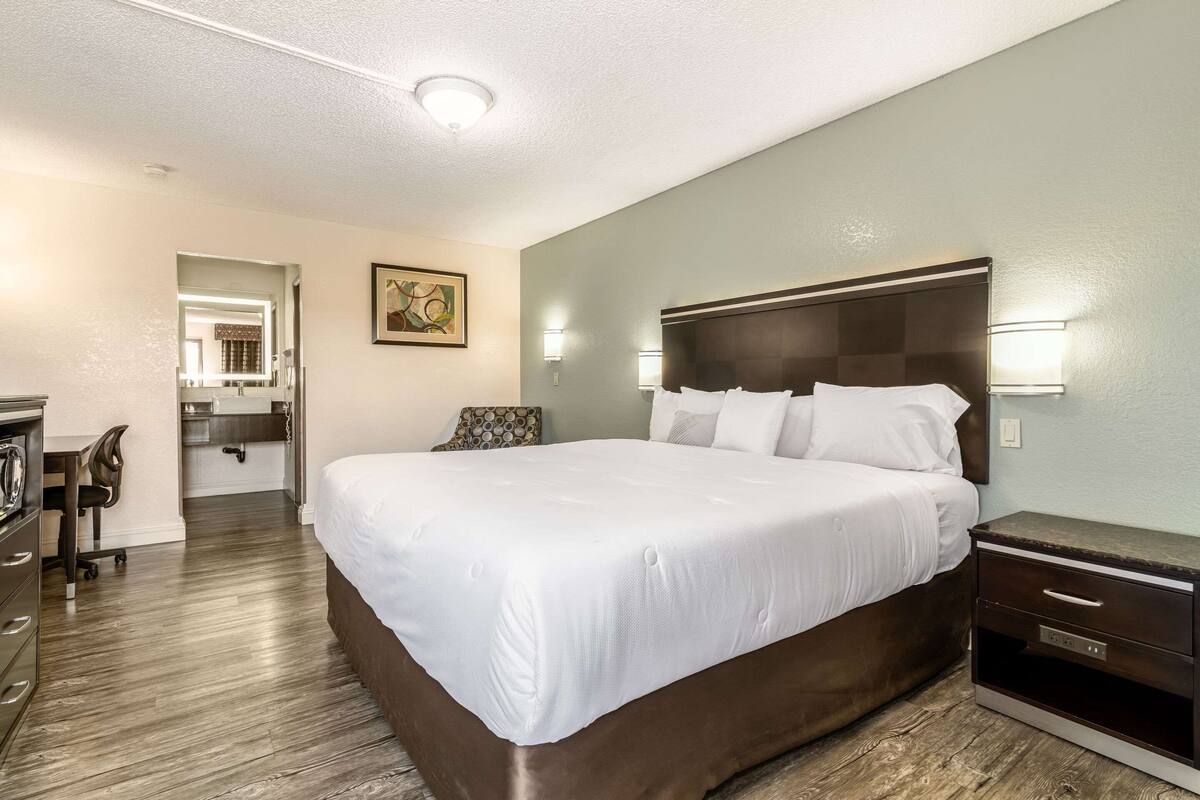 A hotel room with a large bed, a desk with a chair, a dresser, and a nightstand with a lamp.