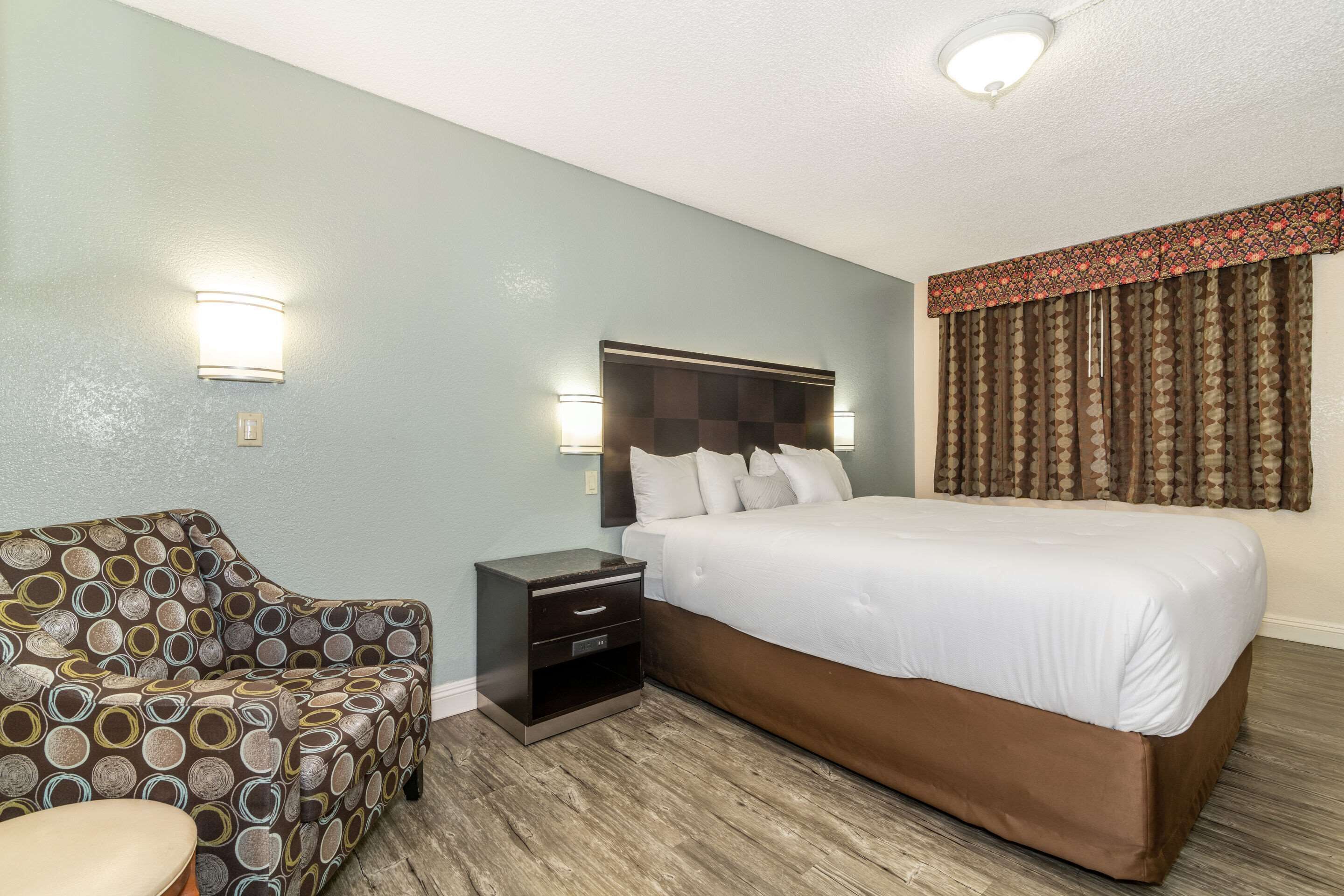 Una habitación de hotel con una cama, una mesita de noche, un sillón estampado y una lámpara fijada a la pared.