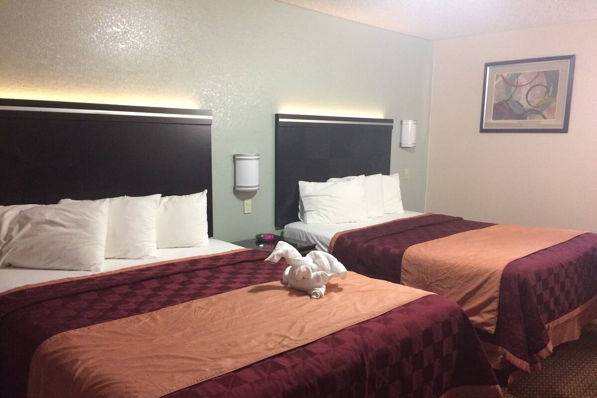 A hotel room with two beds, each with a headboard, white bedding, and a red and beige checkered blanket. There is a white wall-mounted light fixture above the beds and a framed abstract painting on the wall.