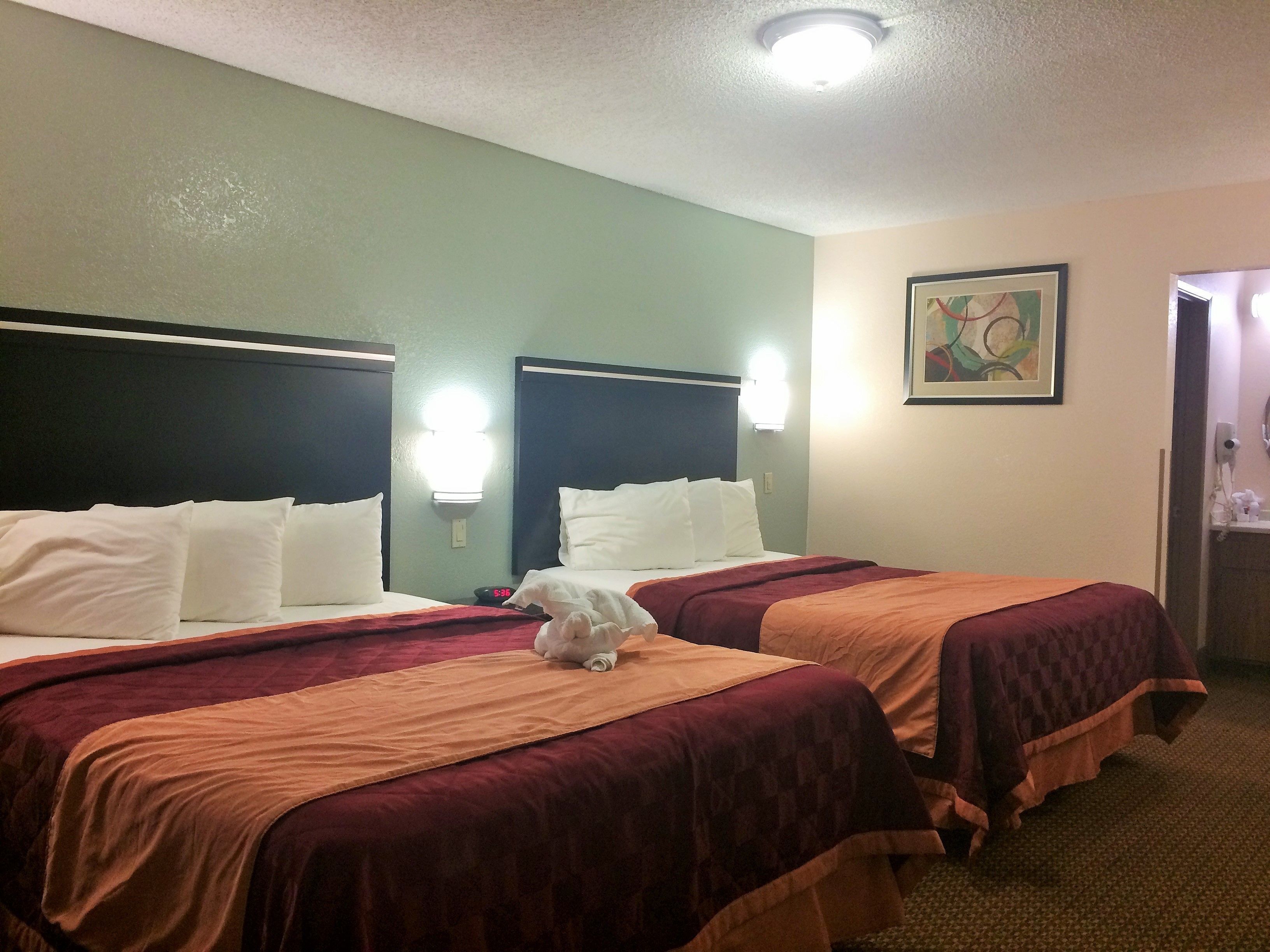 Quarto de hotel com duas camas, cada uma com roupa de cama branca e colchas castanhas. Há um quadro na parede e um relógio na mesa de cabeceira.