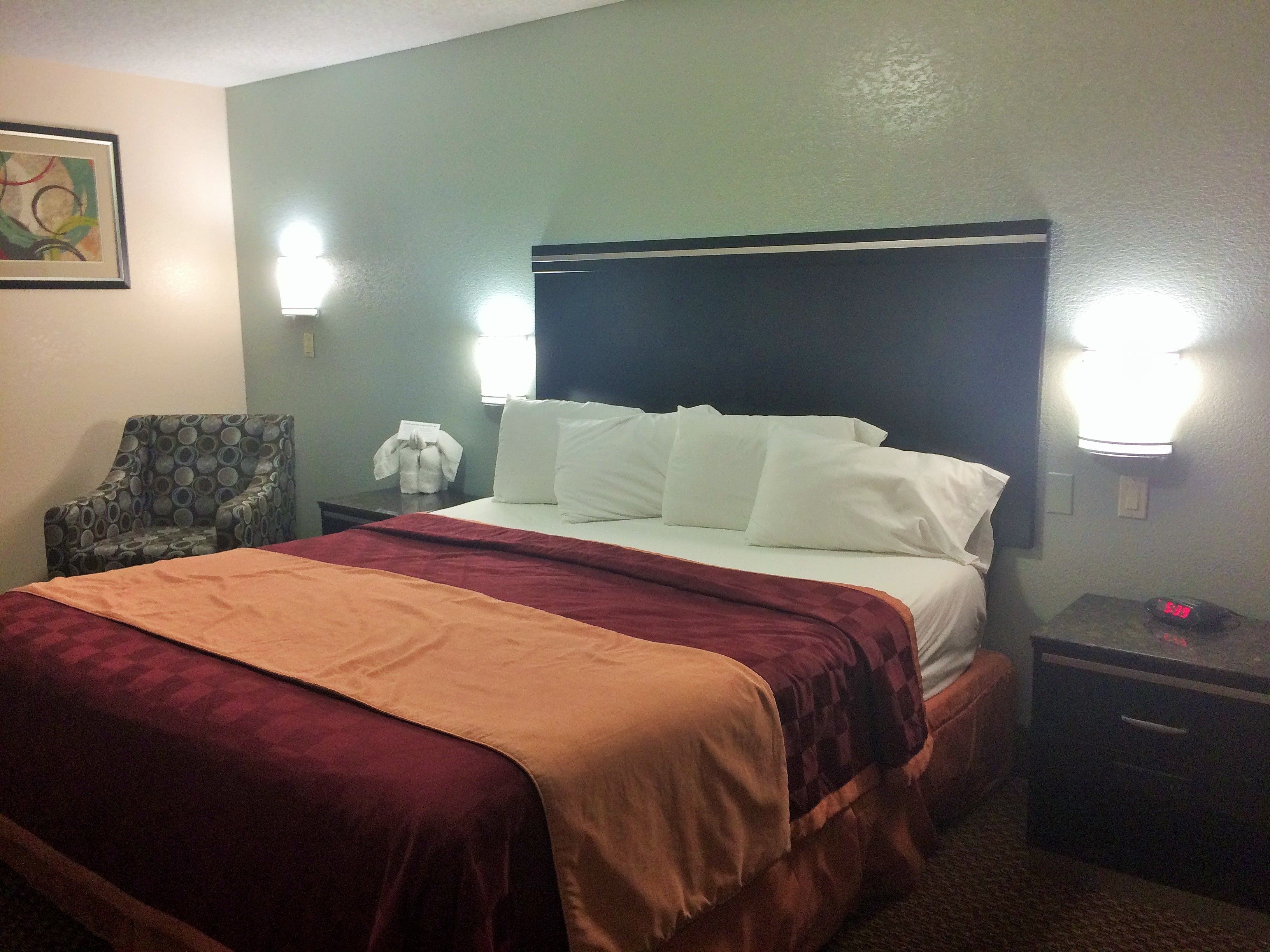 A hotel room with a bed, a chair, a nightstand, and a wall-mounted light fixture.