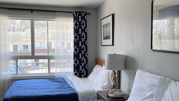 A hotel room with two beds, a window with a view of buildings, a lamp, and a framed picture on the wall. - Knights Inn Midland, ON (Midland)