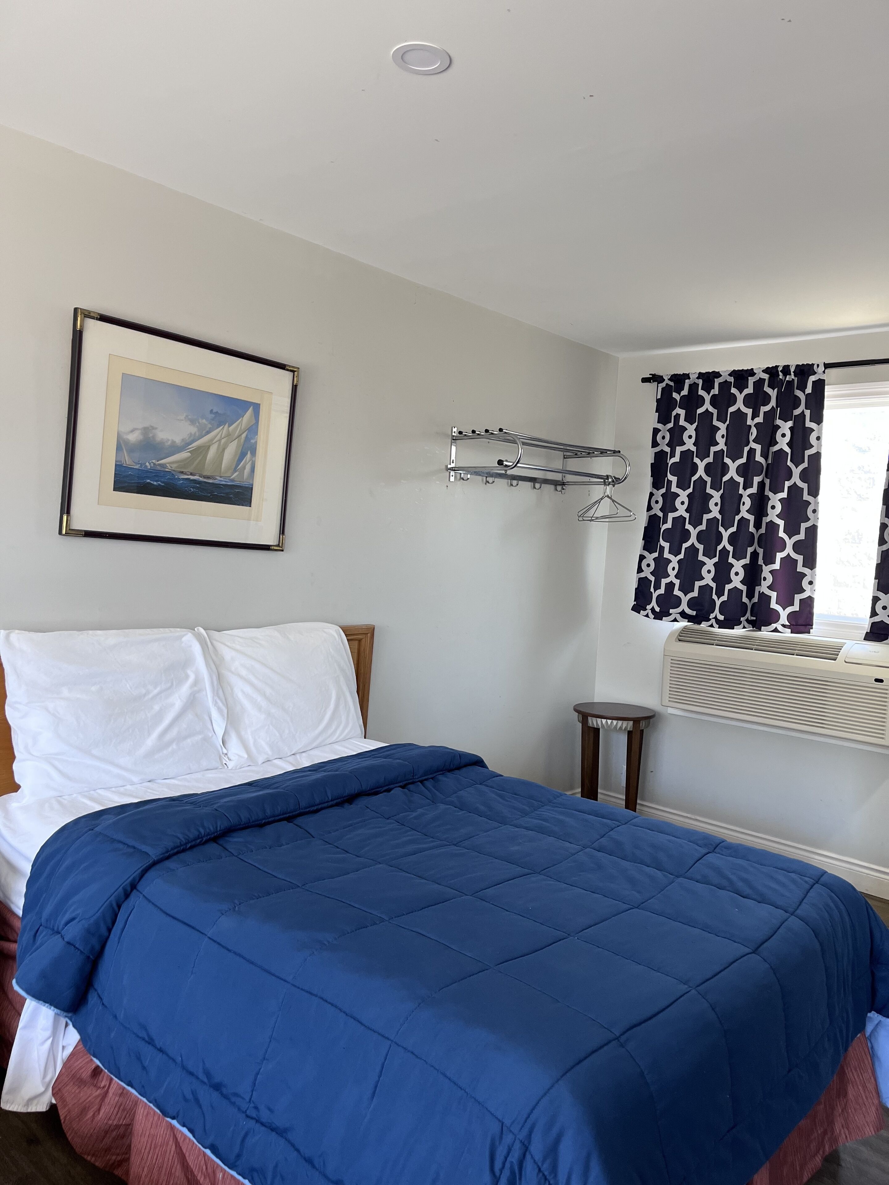 A bedroom with a bed, a framed picture on the wall, a radiator, and a window with patterned curtains.