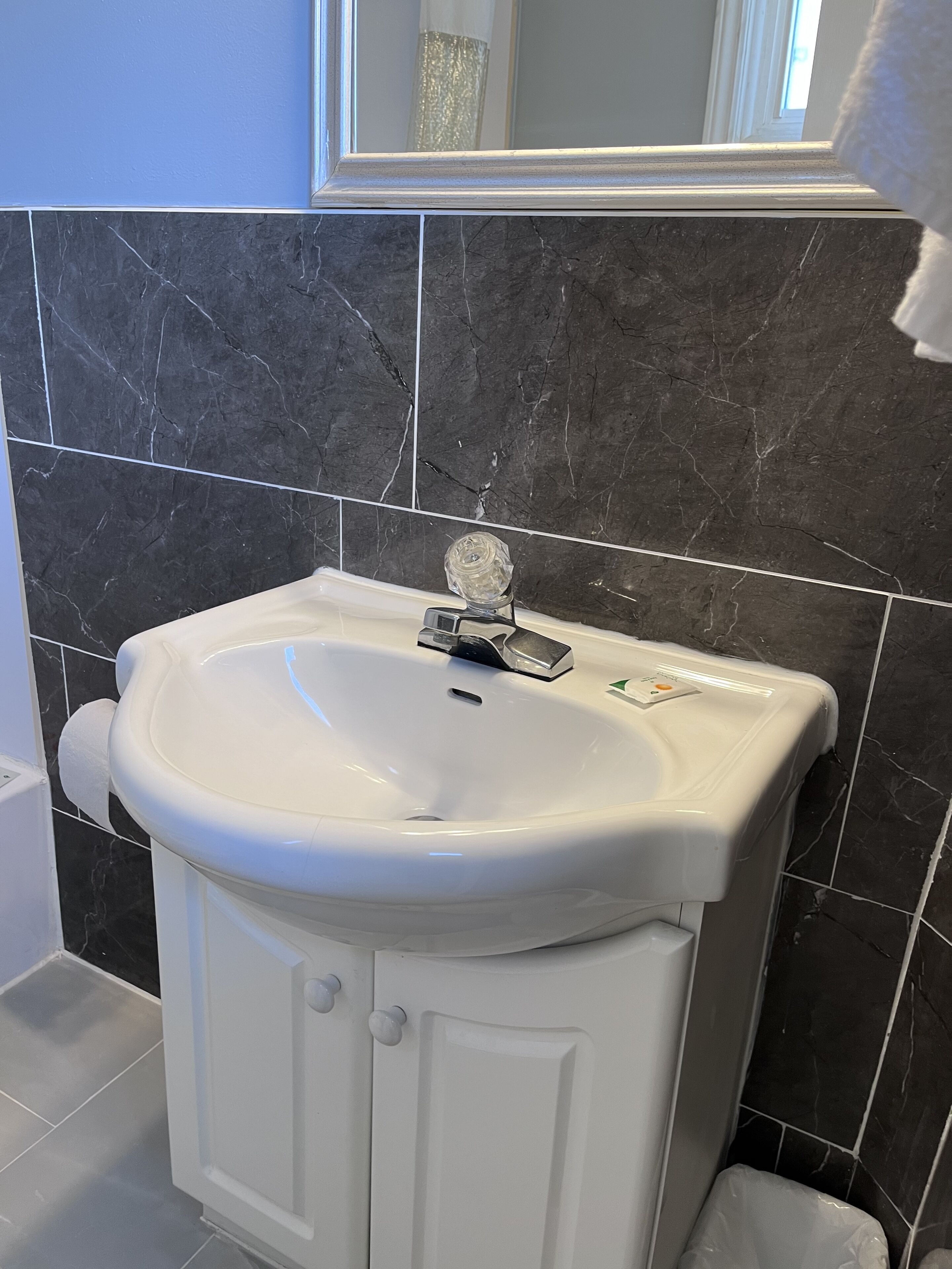 Un baño con un lavamanos pedestal blanco, un espejo encima y baldosas de mármol oscuro en las paredes.