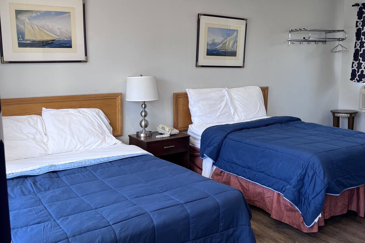 A hotel room with two beds, each with a blue bedspread and white pillows. There are framed paintings on the walls, a bedside table with a lamp, and a window with a curtain.