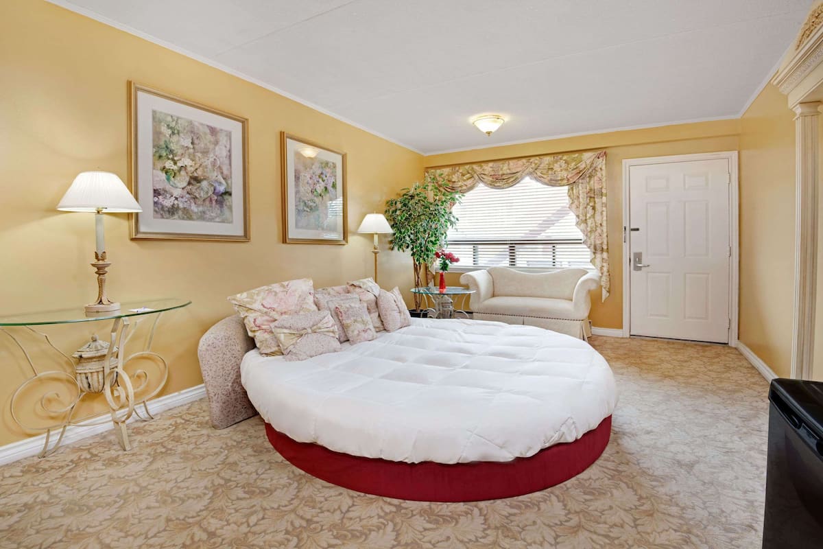 A bedroom with a large bed, a glass table, a chair, and a door.