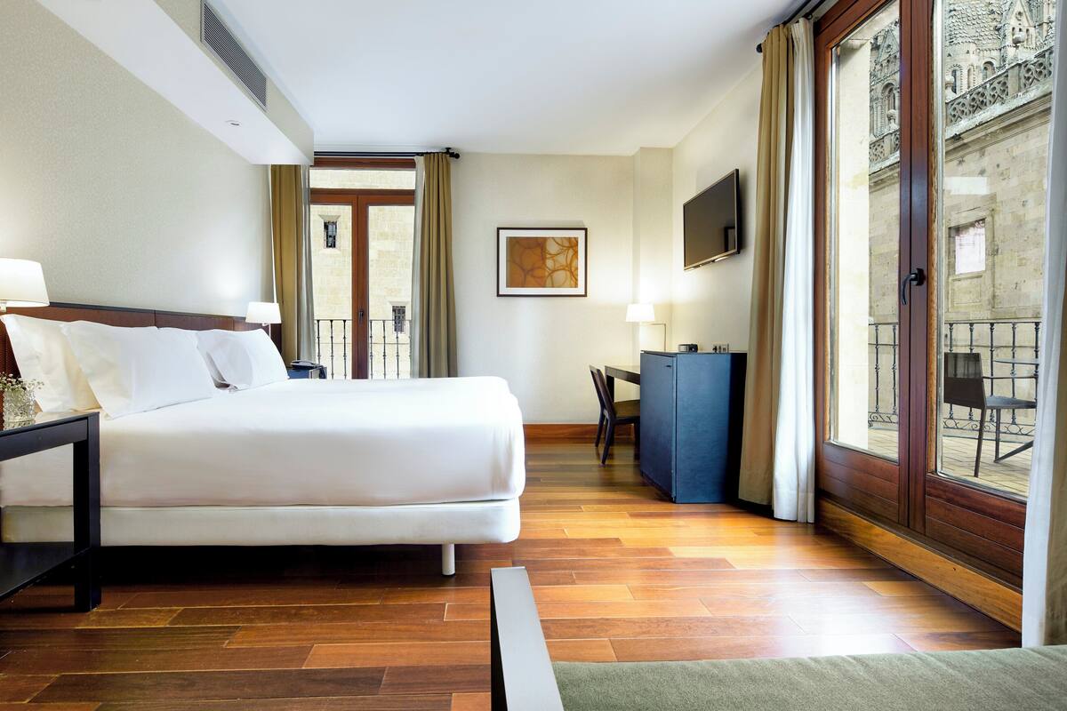 Habitación de hotel con cama, escritorio, silla, televisor y vista a un edificio a través del ventanal.