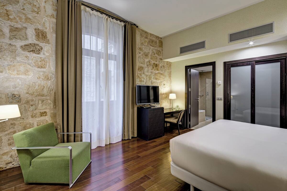 Habitación de hotel con pared de piedra, una cama, un sillón verde, un escritorio con televisor y un baño visible a través de una puerta abierta.