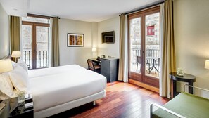 A hotel room with a large bed, a desk, a chair, a small table with a vase, and a balcony with a view of the city.