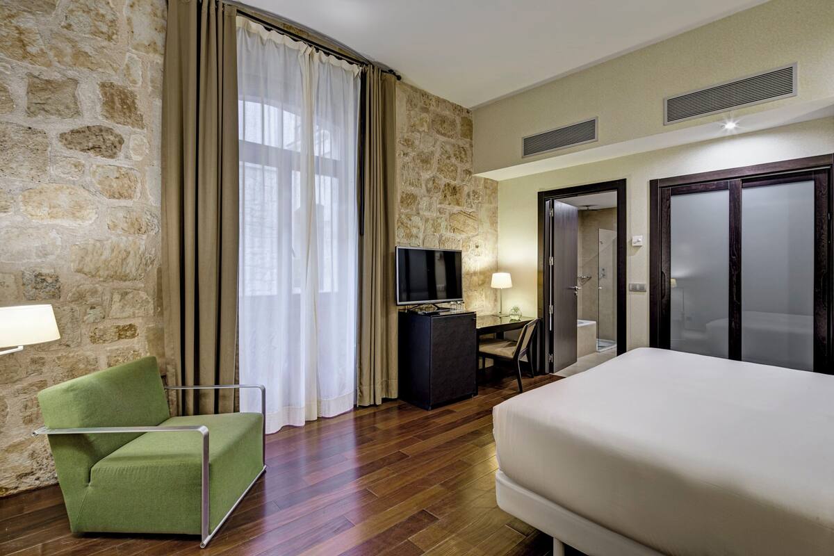 A hotel room with a stone wall, a bed, a green armchair, a desk with a TV, and a bathroom visible through an open door.