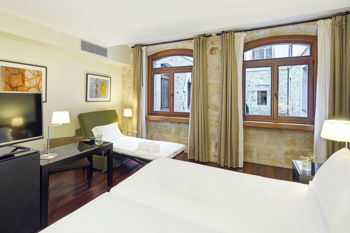 A hotel room with a bed, a desk with a TV, two bedside lamps, and a window with curtains.