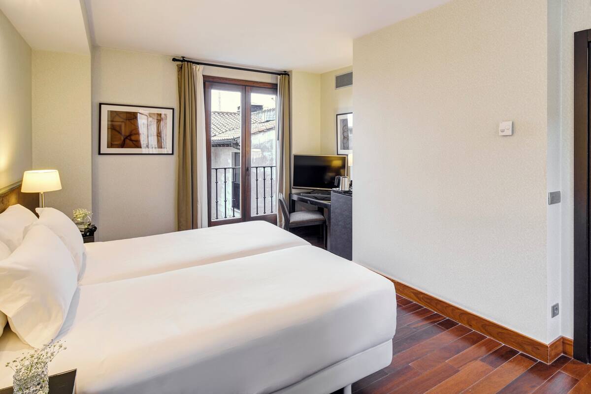 A hotel room with a double bed, a desk with a computer, a TV, and a balcony with a view.