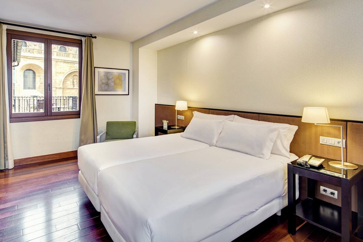 Habitación de hotel con una cama grande, dos mesitas de noche con lámparas, una silla, una ventana con cortinas y vistas a un balcón exterior.