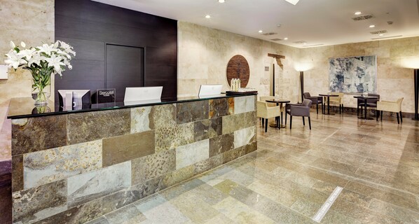 Zona de recepción con pared de piedra, dos mostradores, un jarrón con flores blancas y un comedor con sillas y mesas.