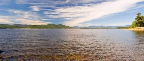 A clear lake with visible rocky bottom and distant mountains under a partly cloudy sky.