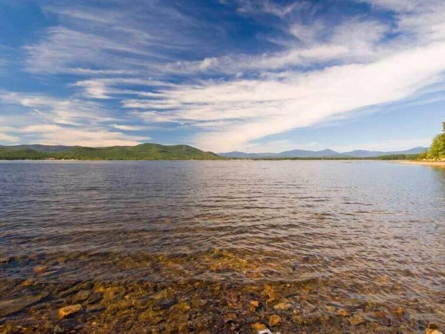 Un lac aux eaux limpides, dont on peut apercevoir le fond rocheux, avec des montagnes lointaines se découpant sur un ciel partiellement nuageux.