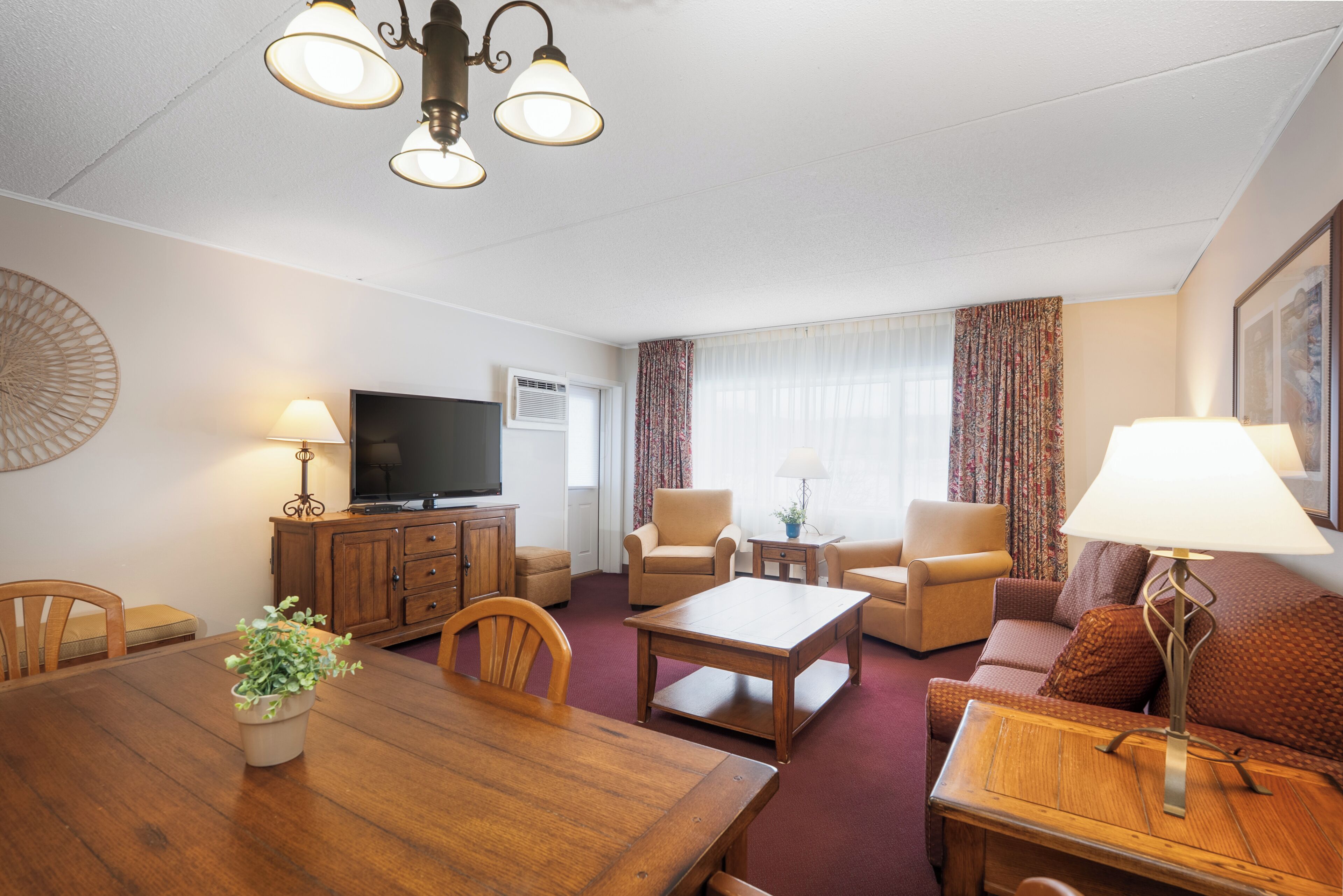 A hotel room with a wooden dining table, two armchairs, a sofa, a TV, and a lamp.