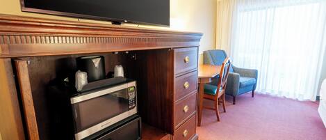Habitación de hotel con una televisión de pantalla plana sobre un mueble de madera, un microondas y una cafetera. Se ve una silla y una mesa con otra silla al fondo.