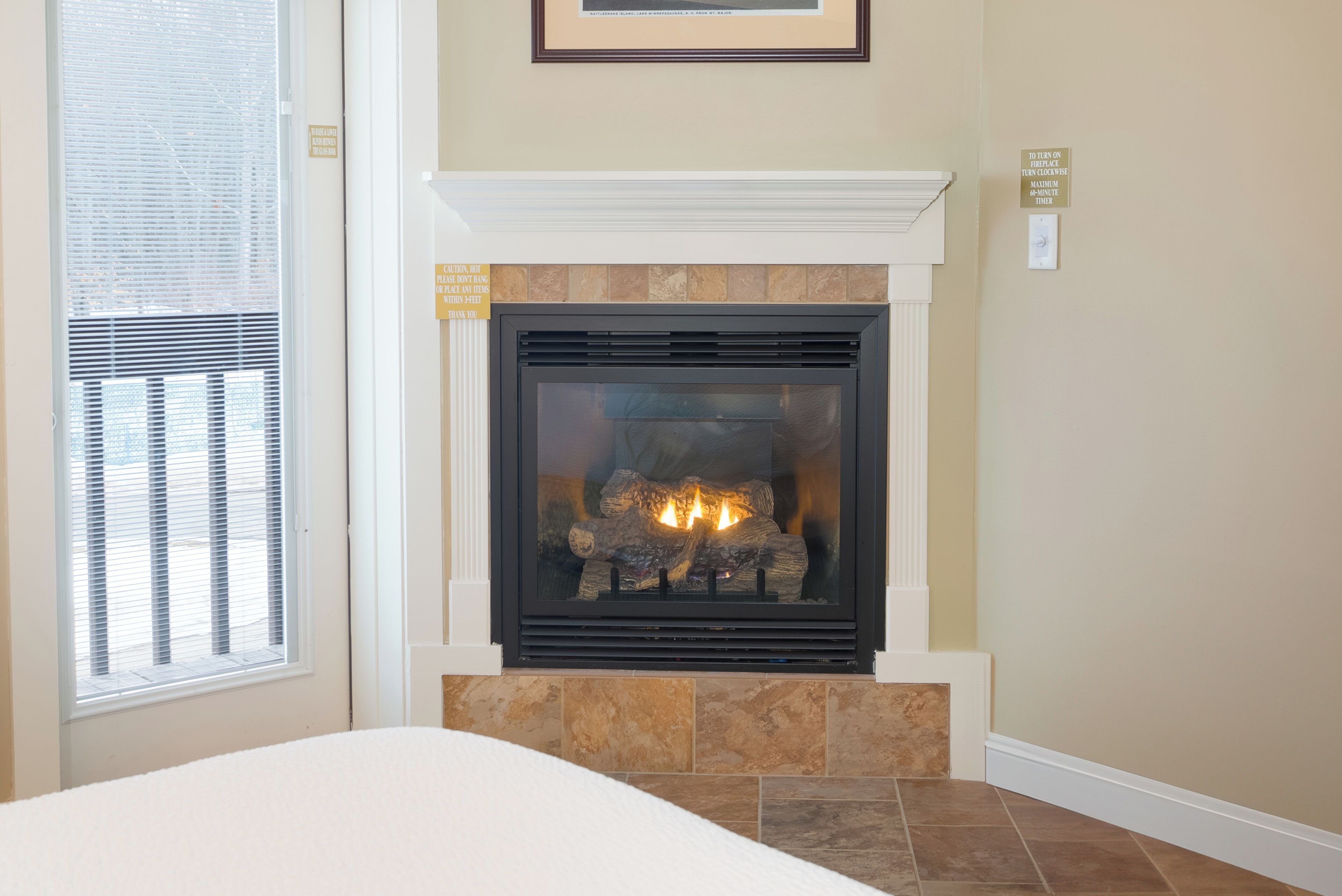 A fireplace with a stone surround, a window with blinds, and a framed picture above the fireplace.