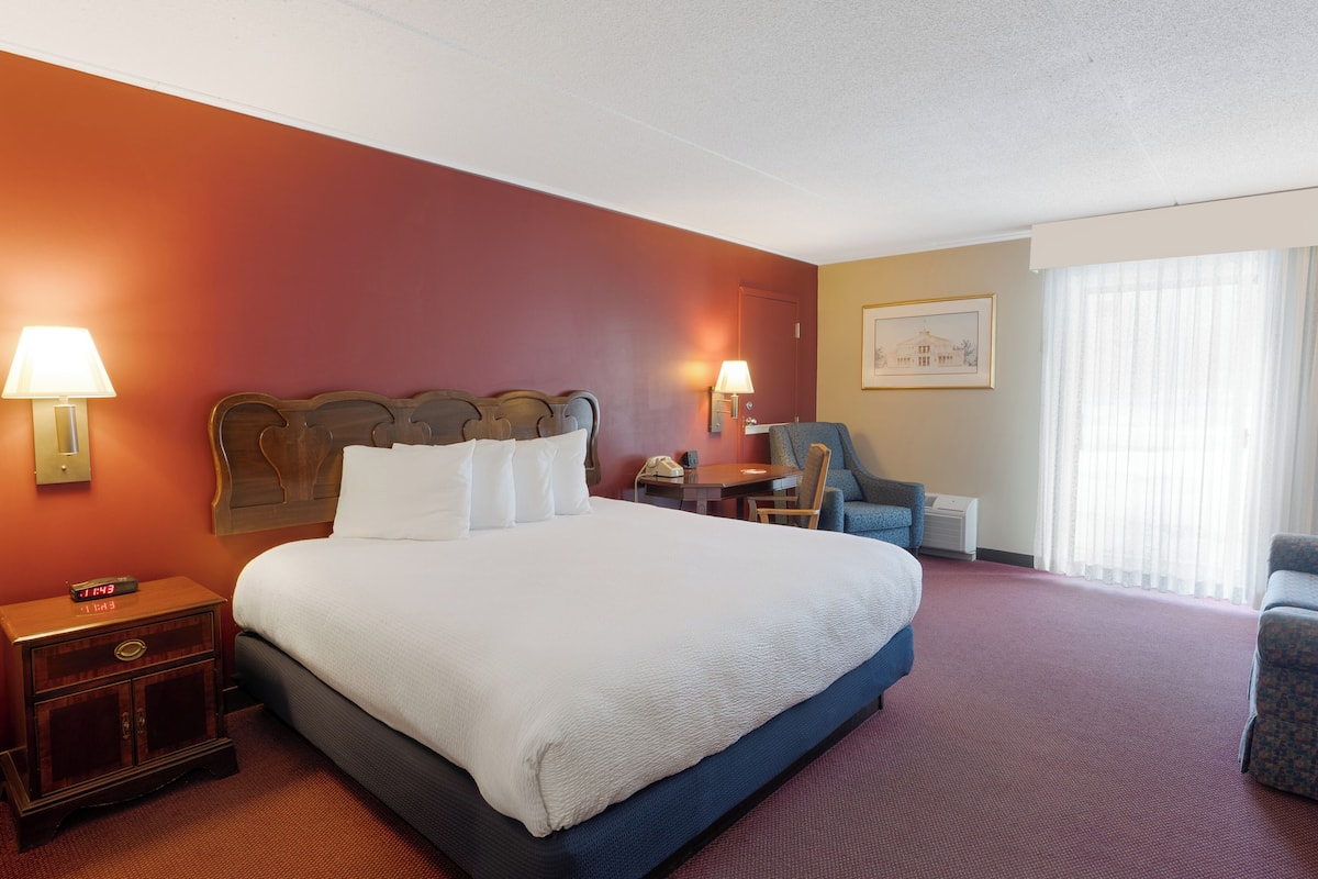 A hotel room with a large bed, two bedside tables with lamps, a desk, a chair, and a large window with curtains.