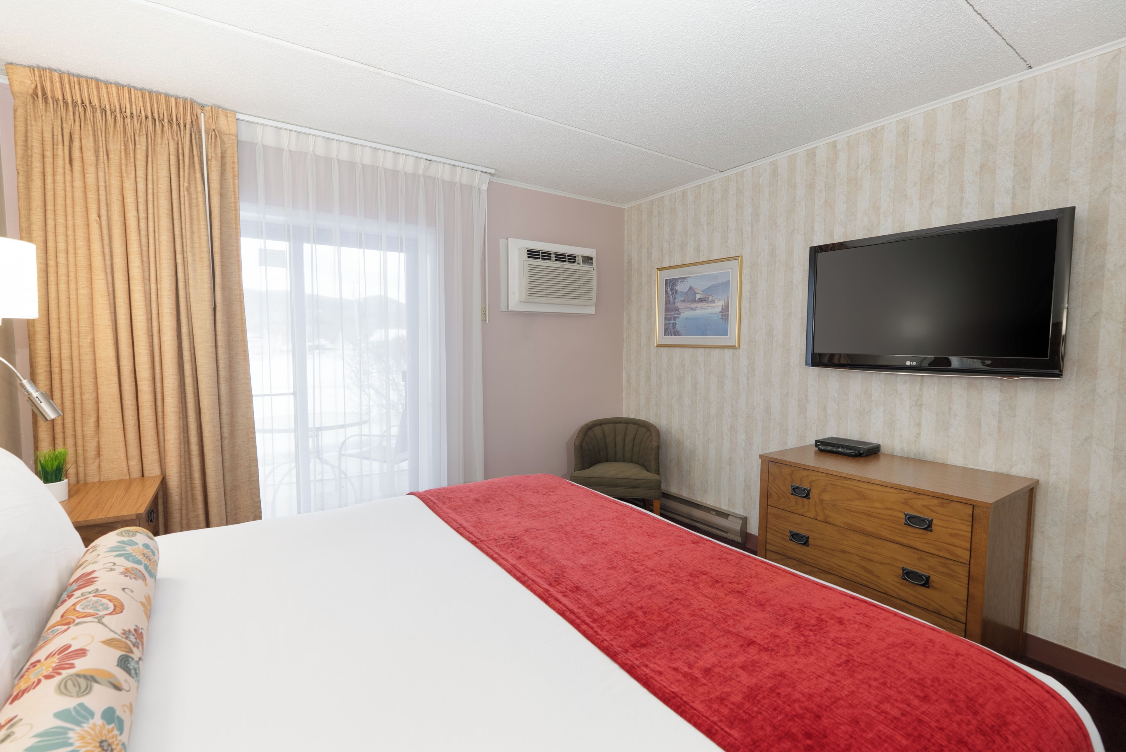 Une chambre d’hôtel avec un lit, une télévision, une chaise, une commode et une fenêtre avec des rideaux.