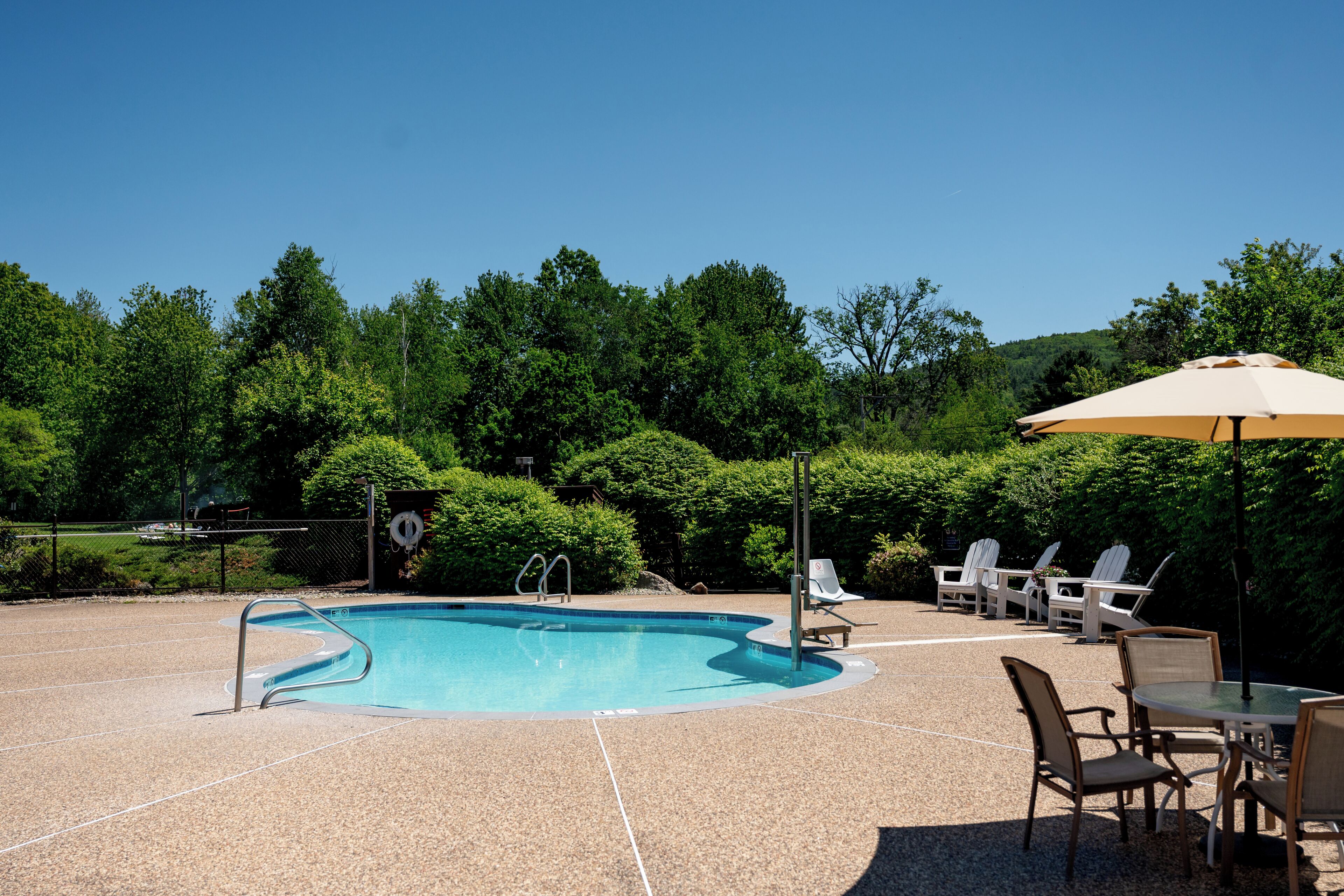 Una piscina con terraza, rodeada de árboles y mobiliario exterior.