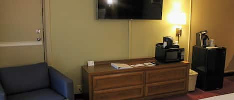 A hotel room with a flat-screen TV mounted on the wall, a wooden dresser with a microwave, a coffee maker, and a lamp. There is a blue armchair and a patterned ottoman.